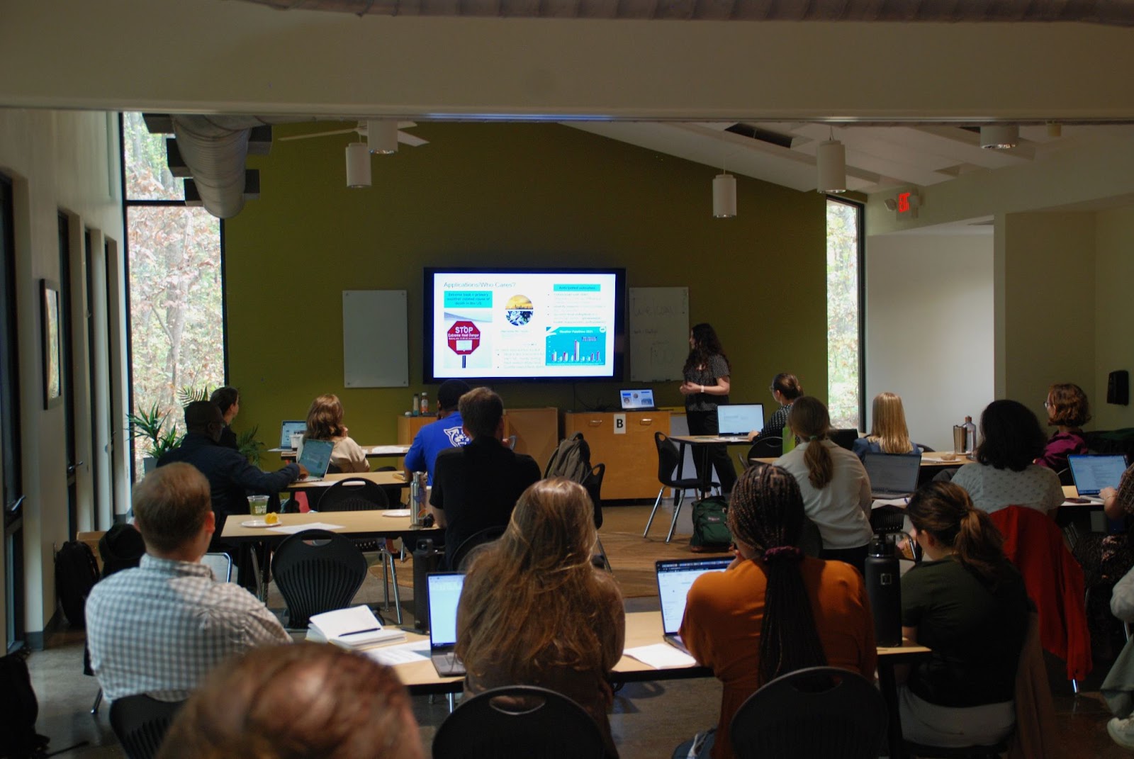 A speaker stands at the front of the room in front of a TV displaying a PowerPoint. Rows of tables fill the room, with several people at each table listening