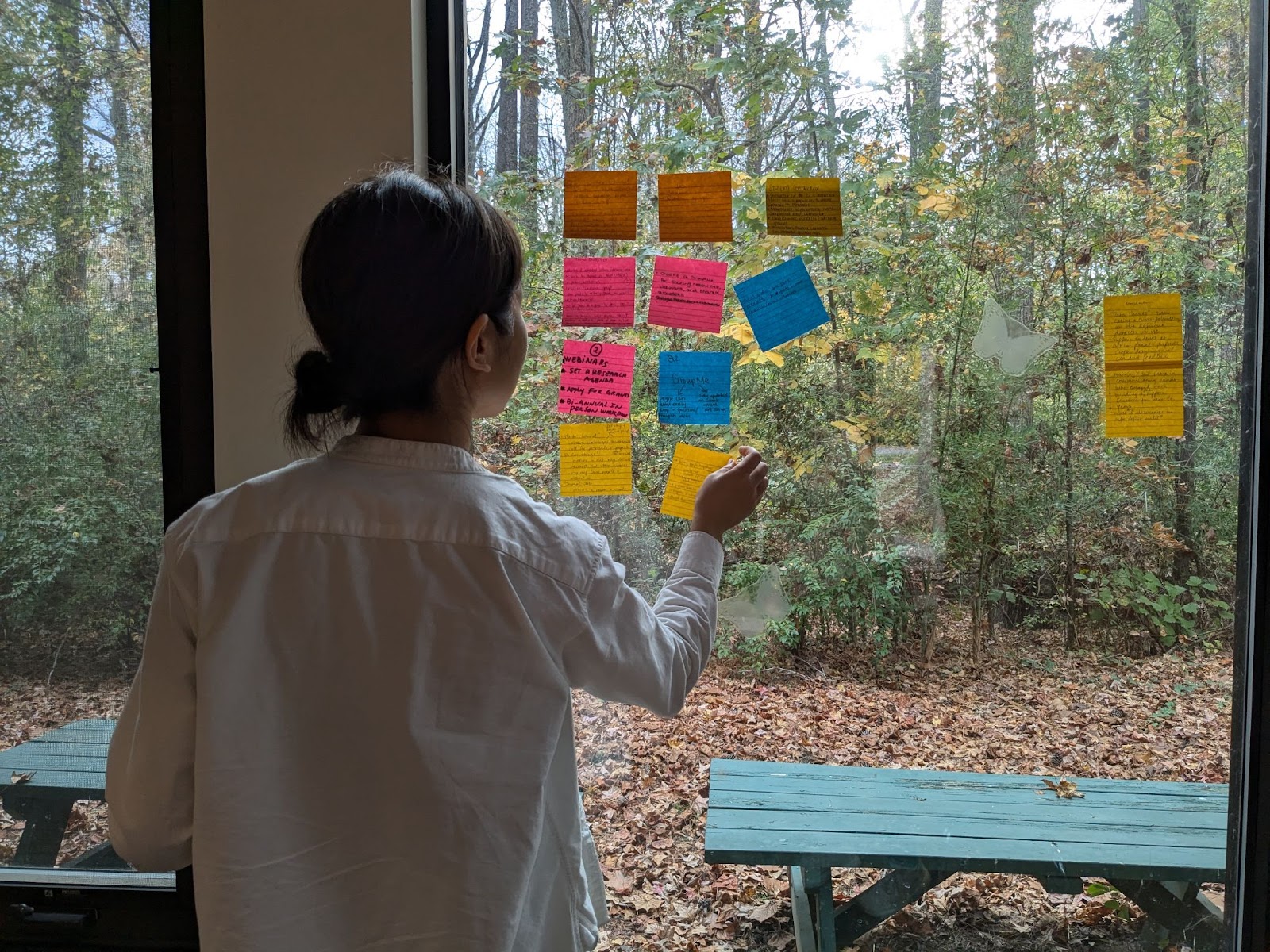 A workshop attendee posts a yellow sticky note on a glass window. The window has 11 other sticky notes already stuck to it, of pink, blue and yellow colors