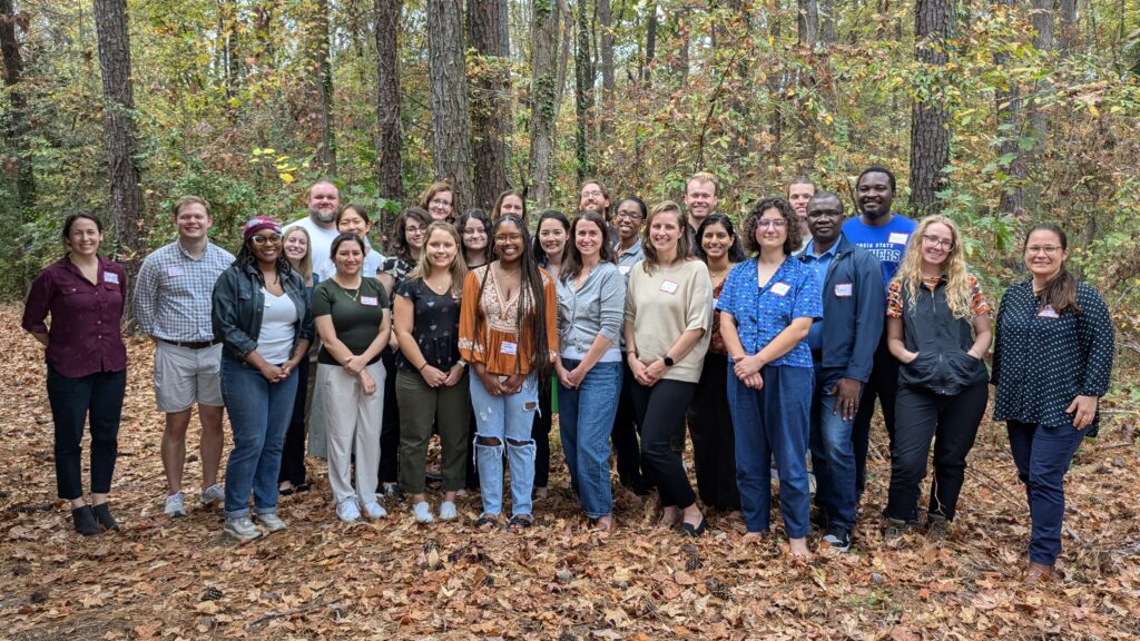 A group of about 25 C3HE workshop attendees pose for a picture outside. There are about three rows of people, standing in front of a wooded background. Leaves cover the ground in varying shades of orange and brown.