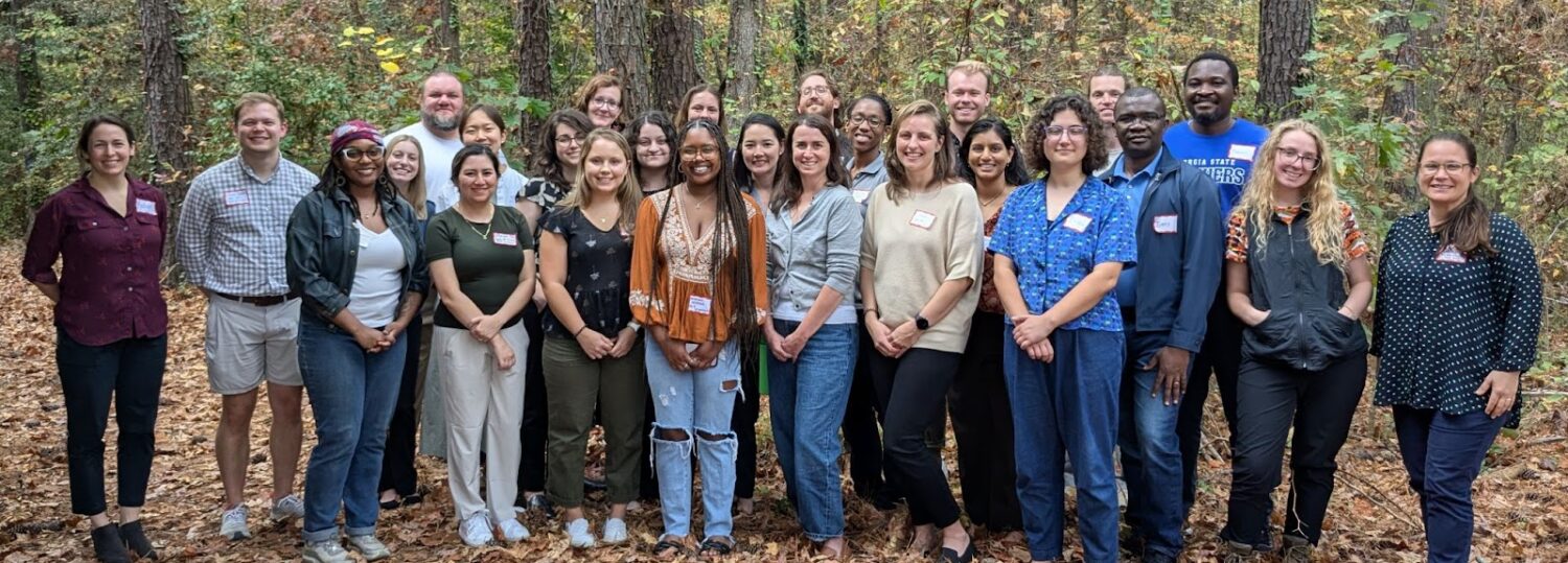 A group of about 25 C3HE workshop attendees pose for a picture outside. There are about three rows of people, standing in front of a wooded background. Leaves cover the ground in varying shades of orange and brown.