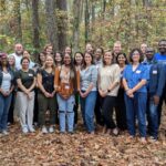 A group of about 25 C3HE workshop attendees pose for a picture outside. There are about three rows of people, standing in front of a wooded background. Leaves cover the ground in varying shades of orange and brown.