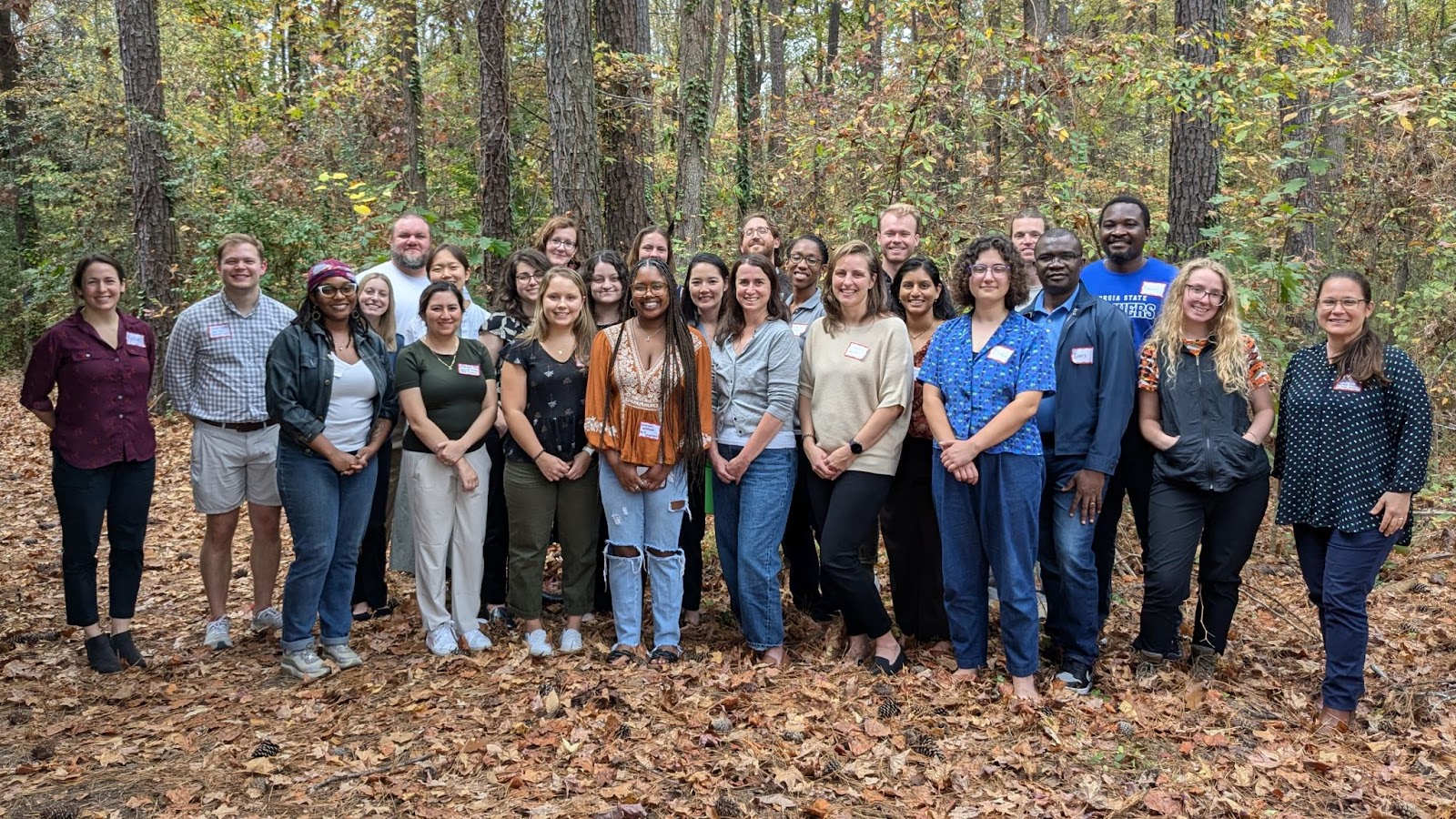 A group of about 25 C3HE workshop attendees pose for a picture outside. There are about three rows of people, standing in front of a wooded background. Leaves cover the ground in varying shades of orange and brown.